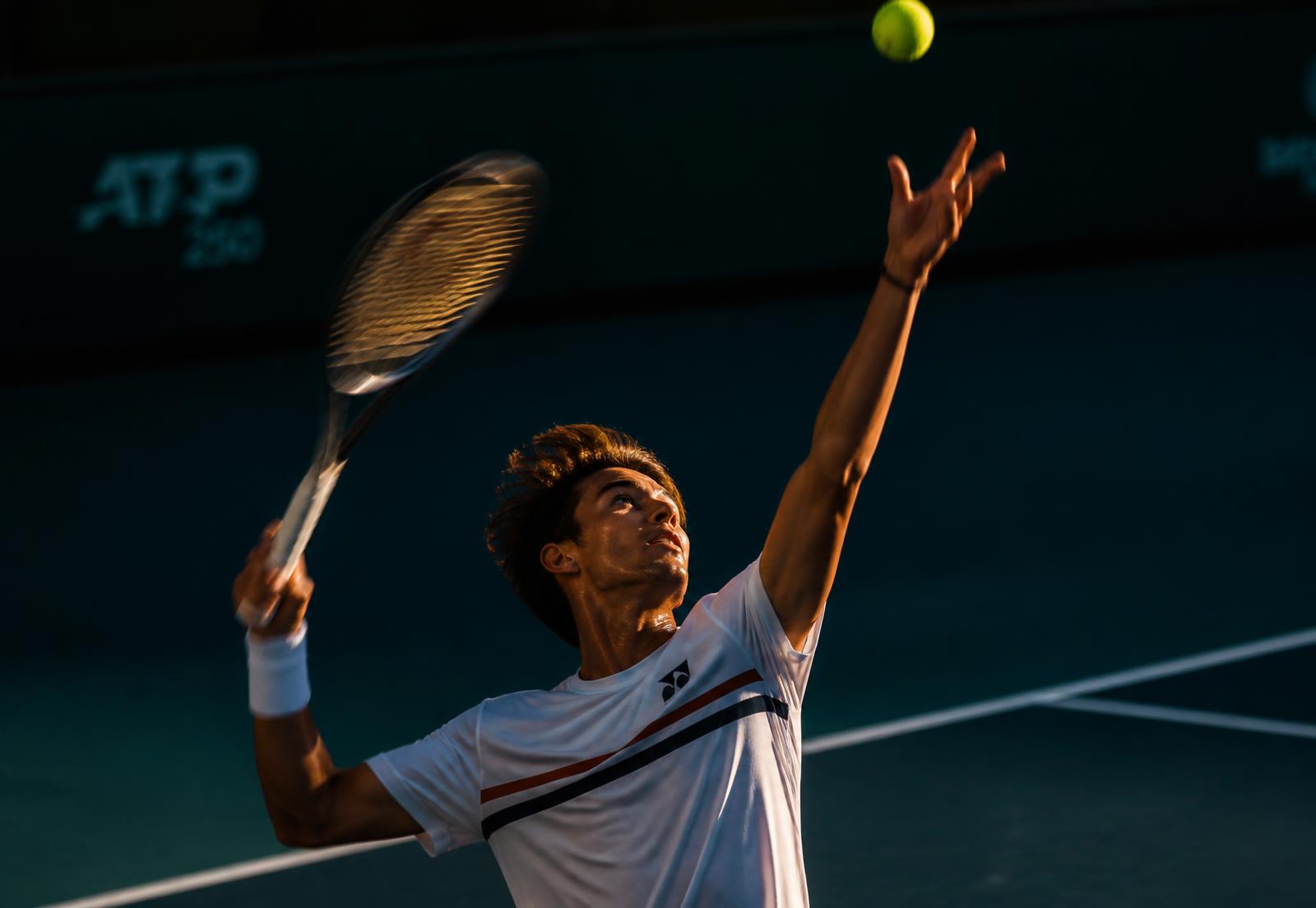 Tennis player mid-serve on a floodlit hard court